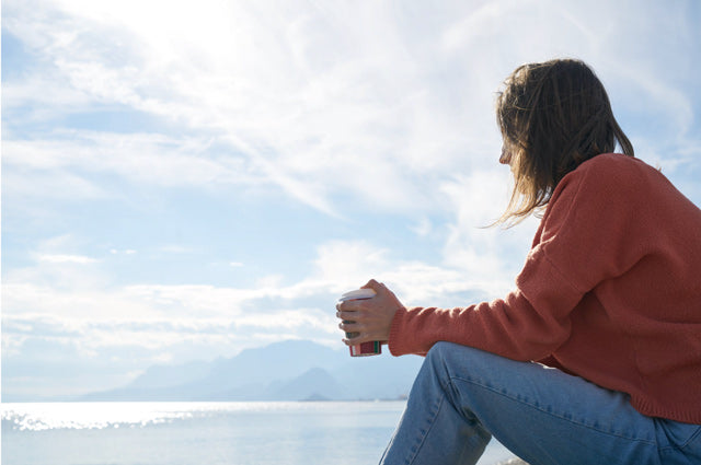 woman in jeans and orange sweater holding coffee looking at body of water